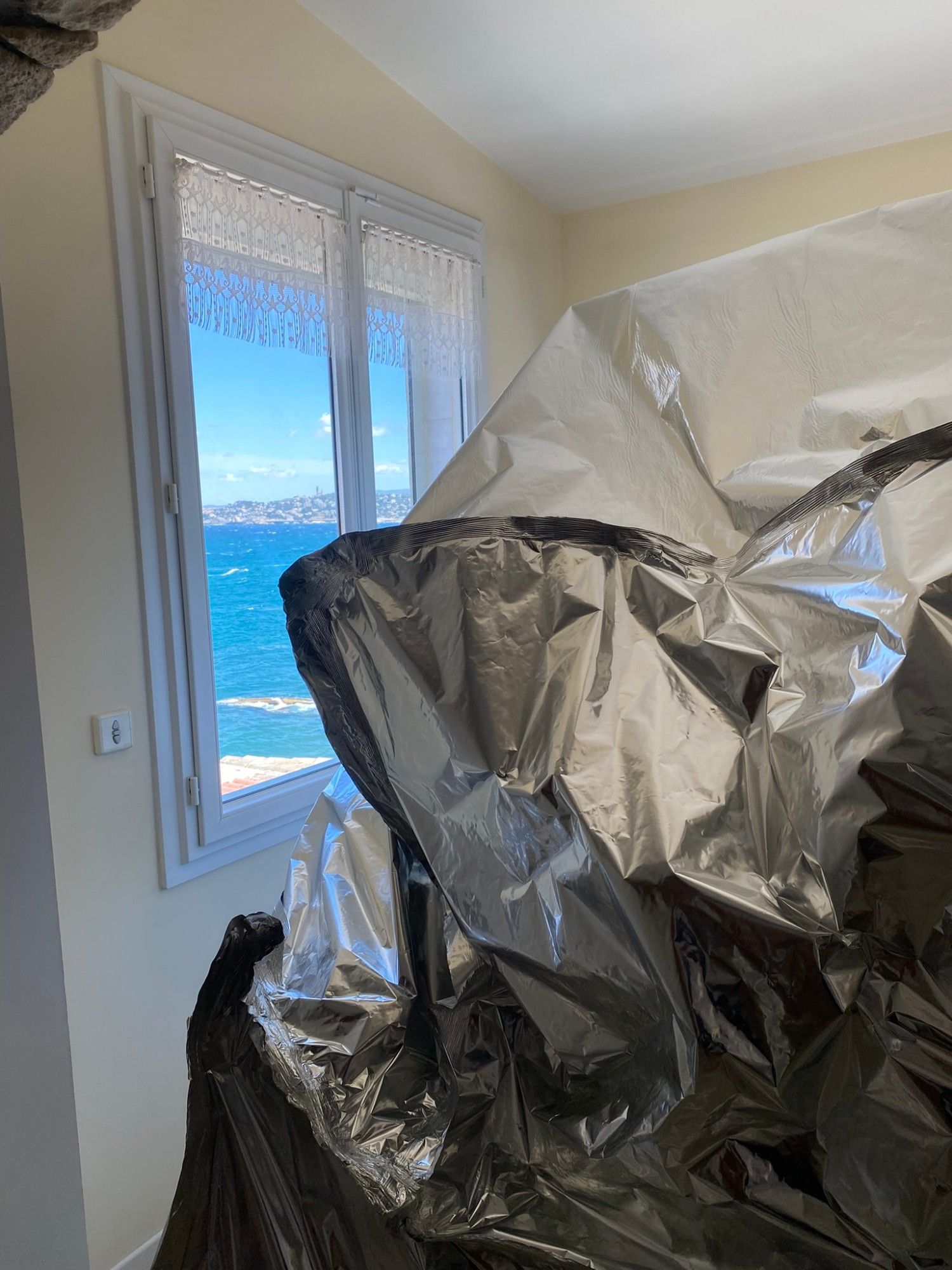 Enveloppe étanche en film aluminium fermée autour de meubles anciens, devant une fenêtre donnant sur la mer
