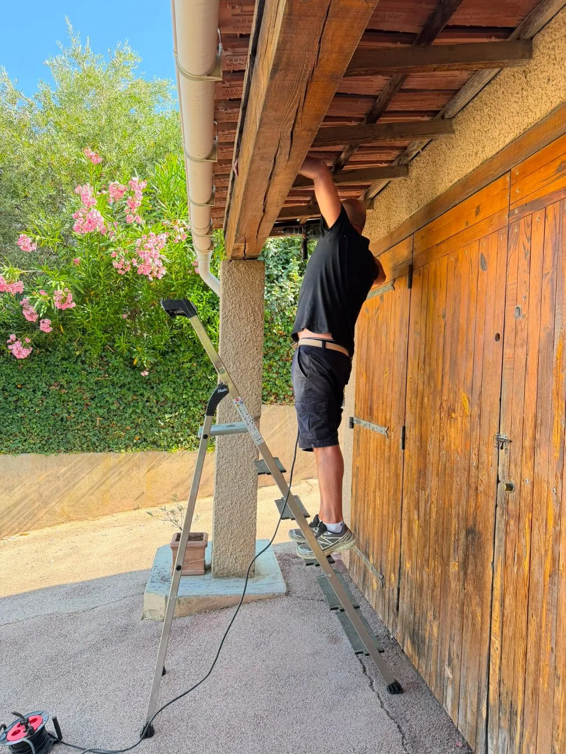 Pascal Sergent sur un escabeau perçant une poutre de terrasse couverte, devant les portes en bois d’un garage