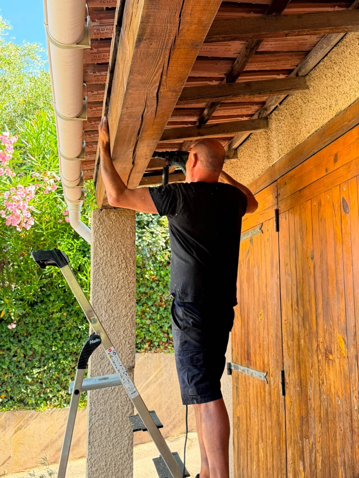Pascal Sergent perçant une grosse poutre de terrasse couverte, sous les tuiles, pour poser les injecteurs