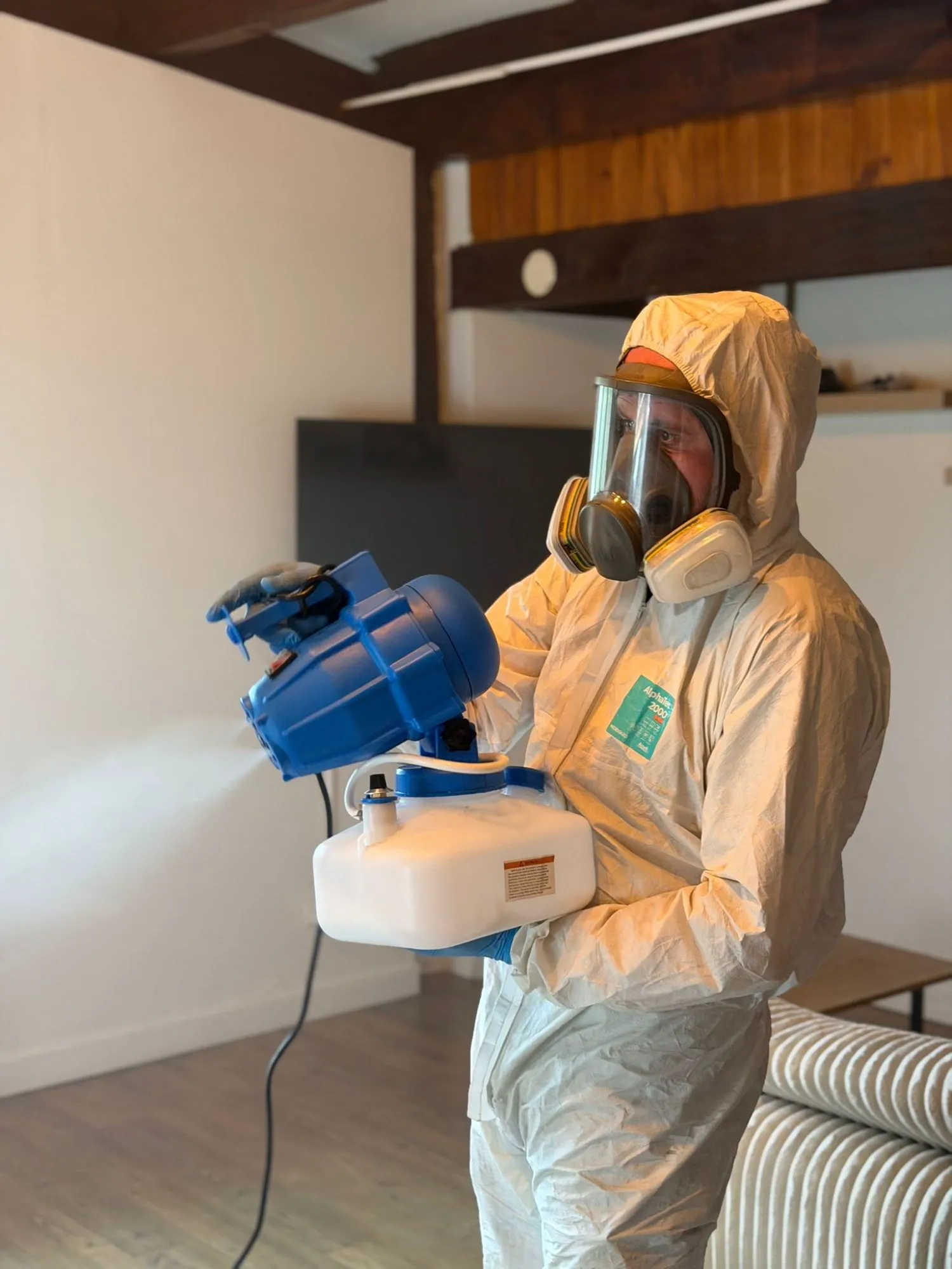 Technicien en combinaison et masque intégral diffusant un brouillard fin avec un nébuliseur électrique dans une pièce sous poutres apparentes