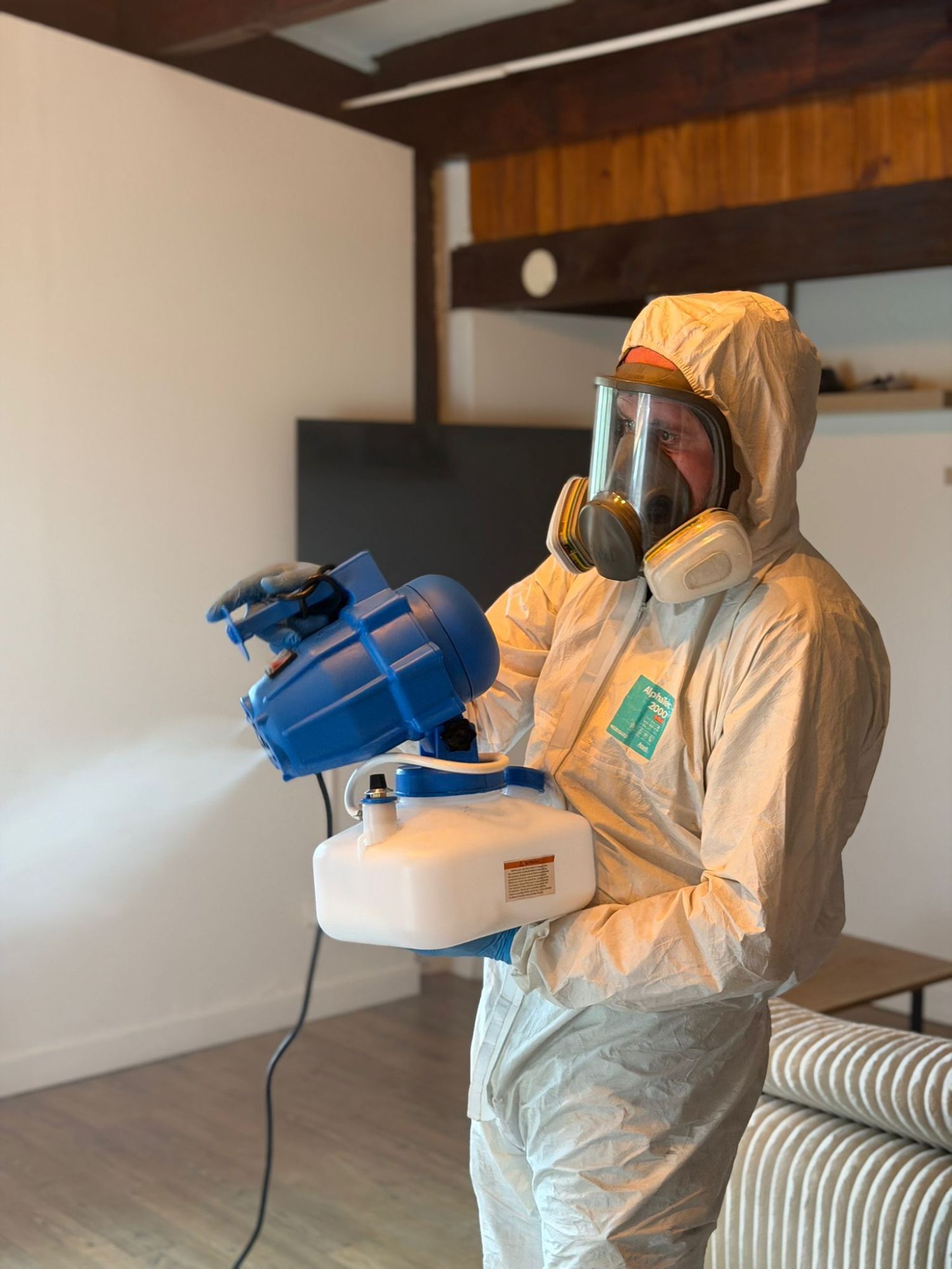 Technicien en combinaison et masque intégral diffusant un brouillard fin avec un nébuliseur électrique dans une pièce sous poutres apparentes