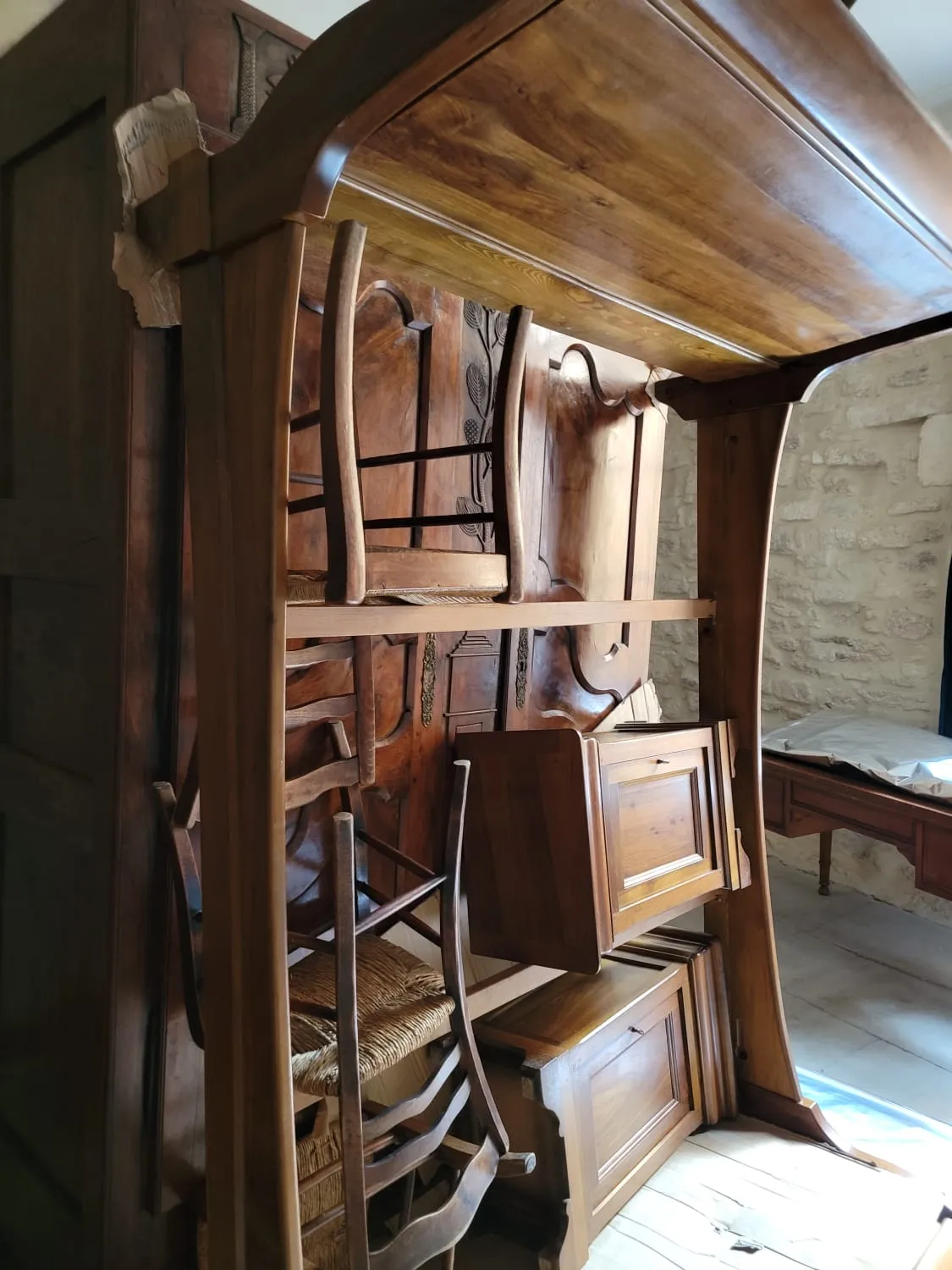 Armoire aux portes sculptées d’un motif végétal, chaises paillées et éléments à abattant rangés sous une grande pièce à fronton courbé