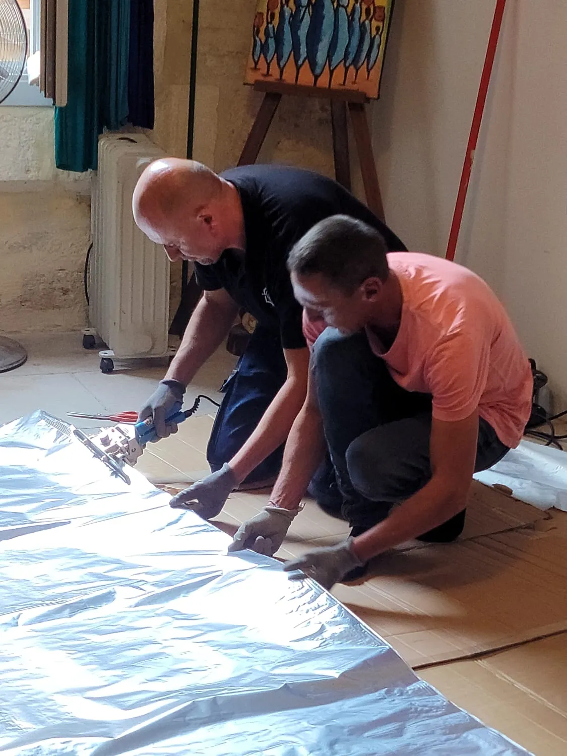 Pascal Sergent et un assistant à genoux, soudant à la soudeuse à main deux lés de film barrière aluminium posés au sol