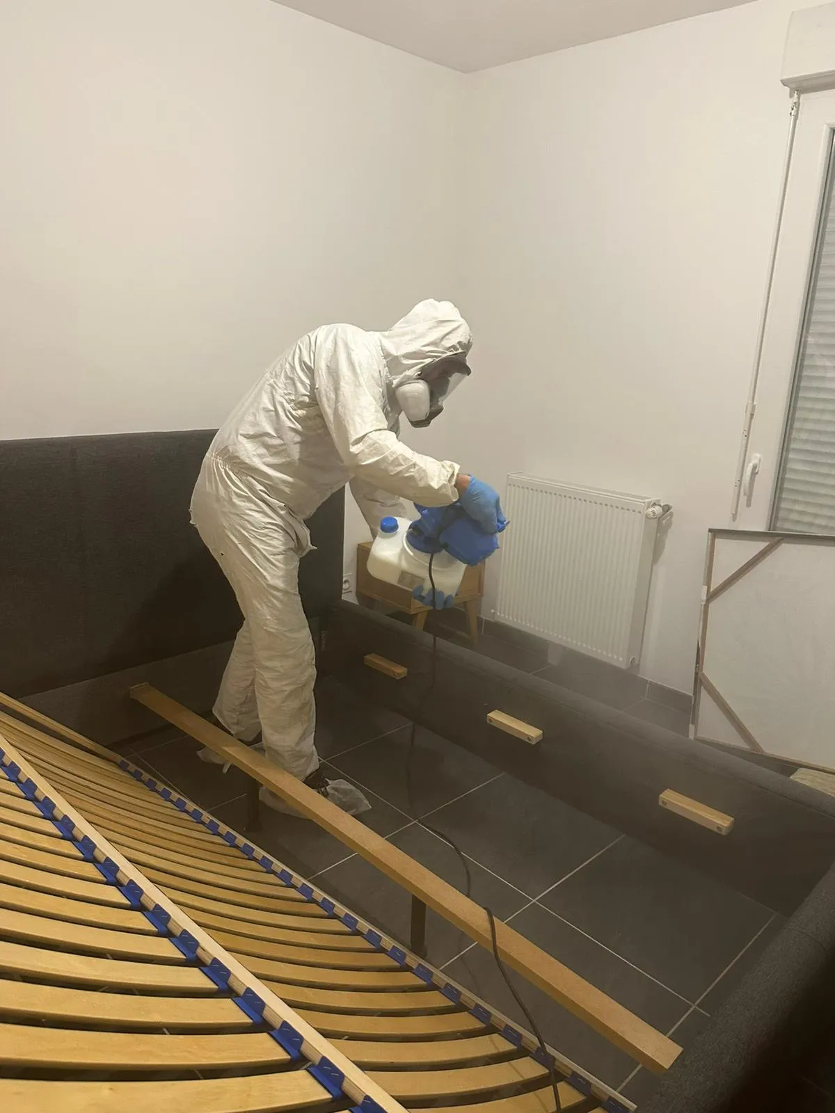 Technicien en combinaison et masque intégral traitant au nébuliseur le cadre d’un lit démonté, sommier à lattes relevé, dans une chambre